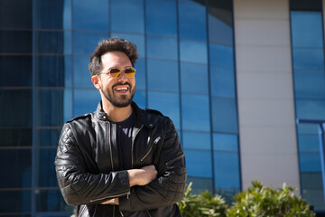 Handsome young man with beard, sunglasses, leather jacket and jeans, arms crossed, smiling happily. Concept beauty, fashion, trend, modern, successful, happiness.