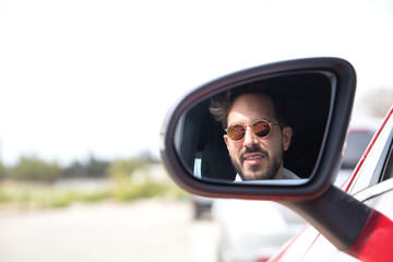 Portrait of handsome young man with beard, sunglasses and white shirt reflected in the rearview mirror of his red sports car. Concept beauty, fashion, trend, luxury, motor, sports, winner, mirror.