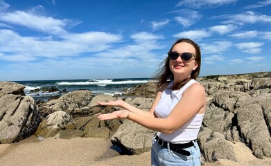 a teenage girl in a white T-shirt shows something with her hands she is wearing black sunglasses and jeans against the backdrop of the ocean she has blond hair. High quality photo