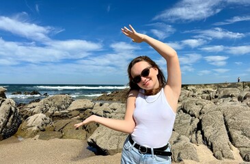 a teenage girl in a white T-shirt shows something with her hands she is wearing black sunglasses and jeans against the backdrop of the ocean she has blond hair. High quality photo
