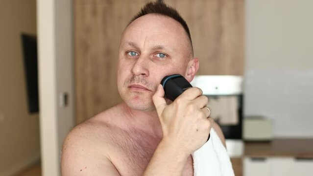 young unshaven man with towel on shoulders shaving with modern electric razor trimer while looking at camera, indoors. Daily routine male self-care. man shaves with an electric razor in the kitchen.