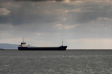 Side view of a cargo ship in the ocean. Dark and moody image with cloudy sky and calm water surface. International shipment and import and export industry. Goods delivery by ocean.