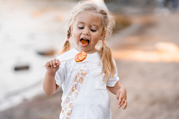 cute little blonde girl having fun in a summer park, eating a colorful lollipop