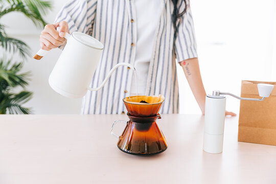 Crop image of professional woman barista with black long hair, hand pouring hot water on ground coffee over the paper filter on a glass dripper. An alternative method is called Dripping.