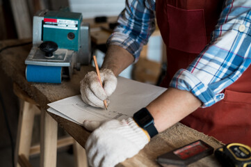 A young Asian carpenter with a brown apron uses a wood sander and tape measure while working on a plank. Carpenter concept working in a wood factory. Selective focus.