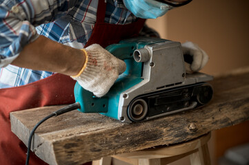 A young Asian carpenter with a brown apron uses a wood sander and tape measure while working on a...