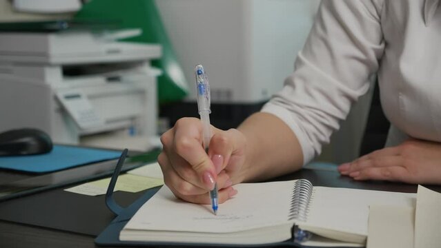 In medical office cabinet young female doctor in open white lab coat with rolled up sleeves sits at table with documents on desk writing cursive words in notebook with blue ink pen, slow motion pan.