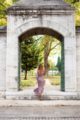 Fototapeta na wymiar Beautiful young woman with long hair in maxi floral dress entering the old stone garden gates, back view. Tourist girl enjoys traveling the old town. Woman have summer vacations go for a walk 