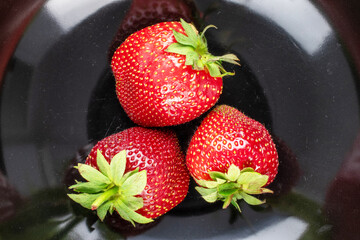 Three berries of ripe organic strawberries on a black ceramic dish, close-up, top view.