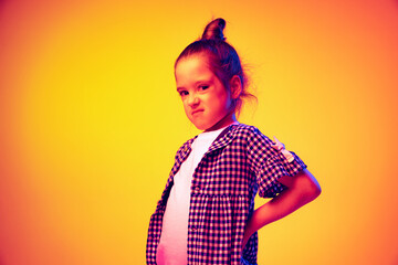 Annoyed little girl, kid posing isolated on yellow red color background in neon light. Concept of children emotions, fashion, beauty, school and ad concept