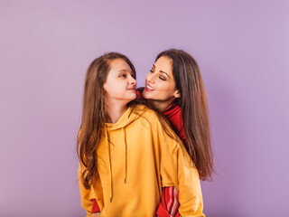 Happy smiling beautiful young mother and cute daughter posing in fashion trendy bright colorful hoodie together on purple studio background with empty copy space. Happy