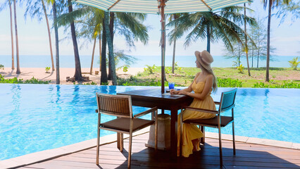 Fototapeta na wymiar Luxury vacation in modern hotel resort. Traveler woman in dress and hat sitting in cafe near swimming pool, drinking welcome refreshment drink and enjoy tropical view with sea and palm trees