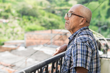 close-up of brown-skinned elderly latino man standing on the balcony of his house looking up at the...