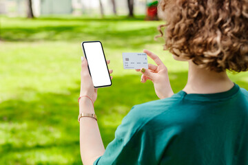 Redhead woman wearing casual clothes hands holding credit card and phone with mockup white blank display, empty screen for shopping or banking app. Mobile app tech concept, over shoulder closeup view.