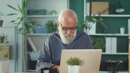 Concentrated Elderly Businessman Working Online While Sitting at His Desk in Bright Room.Tired Man With a Cup Of Tea Thoughtfully Typing on the Keyboard. Remote Work Concept And Retirement Concept.
