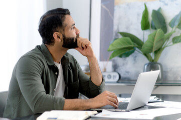 Pensive successful mixed race man, office worker, sitting in a modern office, distracted from work...