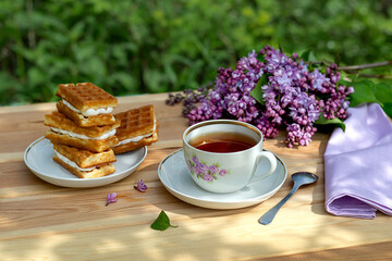 Tea party with Viennese waffles and a branch of lilac on a wooden table against the background of the garden on a sunny summer day.