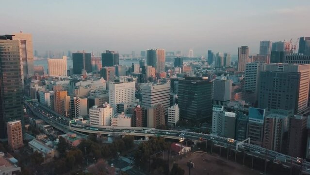 Drone panning up from above the Tokyo city skyline at sunset over a bustling highway bridge full of traffic, tall skyscrapers and orange light reflections with hazy skies