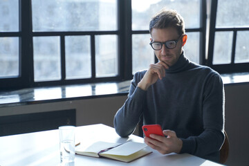 Young german businessman reading internet news or checking email on smartphone while sitting at desk