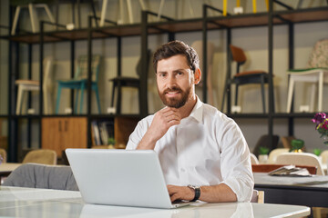 Front view of handsome boy with beard sitting at table working in furniture store. Fashionable...