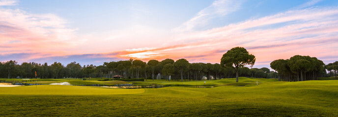 Panoramic view of beautiful golf course with pines at sunset. Golf field with fairway, green, hole,...