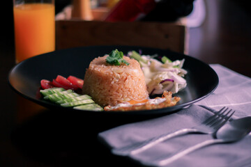 Fried rice, fried egg, cucumber, sliced tomato, green salad and Fried chicken placed on a black plate as a garnish.