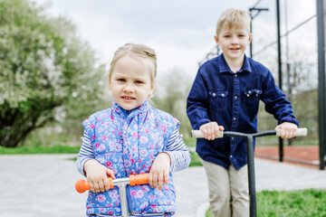 Little blond boy and girl ride kick scooters. Children play in playground on sunny spring day.