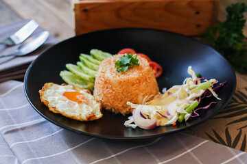Fried rice, fried egg, cucumber, sliced tomato, green salad and Fried chicken placed on a black plate as a garnish.