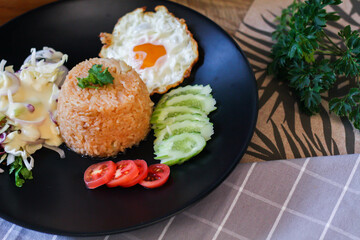 Fried rice, fried egg, cucumber, sliced tomato, green salad and Fried chicken placed on a black plate as a garnish.