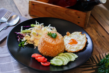 Fried rice, fried egg, cucumber, sliced tomato, green salad and Fried chicken placed on a black plate as a garnish.