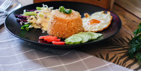 Fried rice, fried egg, cucumber, sliced tomato, green salad and Fried chicken placed on a black plate as a garnish.