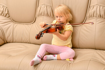 A little girl with a violin in her hands sits on a leather sofa.