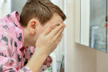 A young man washes his face with water from the tap in the washbasin and wipes his face with his...