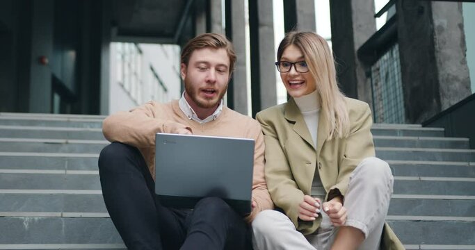 Colleagues caucasian woman and man sitting on the stairs outdoors looking in laptop successful win. Young businesspeople winner making victory gesture. Concept of success.