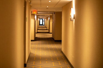  A long narrow hallway of a hotel with a red exit sign light sconce lights and cream colored walls....