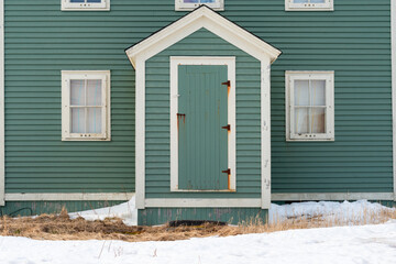 The exterior facade of a vintage wooden building. There's a green single shutter door with rusty hinges, white trim, and wood boards. There are multiple small four pane glass windows with white trim.