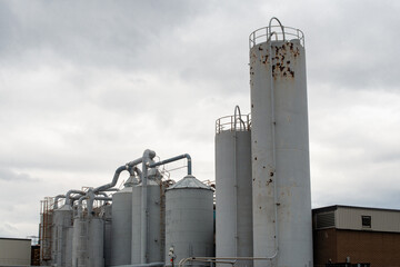 Multiple grey silos of various sizes and heights. The tall round metal silos contain plastic materials, pulp fiber and paint. There's a brick building next to the manufacturing plant with a cloudy sky
