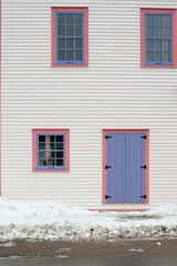The exterior wall of a white wooden cape cod clapboard siding house with a purple panel door and vintage glass window, black metal hinges, pink trim around the door, and white snow on the step. 