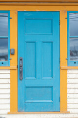 A colorful teal blue color wooden panel door with yellow decorative trim on a white exterior wall. There's a long brass door handle on the wood door and a glass window on both sides of the door. 
