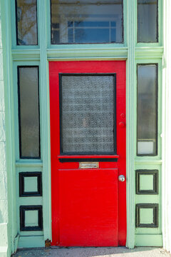 The exterior facade of a vintage shop with an orange wooden door with a glass window, mailslot and door handle. The door is on a lime green building with black trim. There's a transom window over door