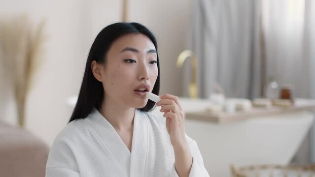 Beauty care at home. Young beautiful asian lady applying moisturizing lipstick on lips and preening her hair at bathroom