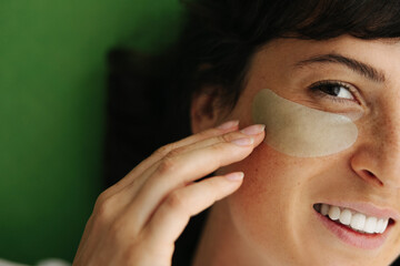 Close-up of pretty young caucasian lady doing facial skin care procedures with hyaluronic patches. Brunette smiling looking at camera on green background.