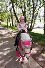 Obraz premium Little girl riding a pony horse in park. Child dressed in pink jacket and hat on horse.