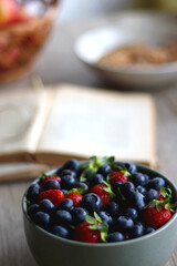 Bowl of strawberries and blueberries, open books with reading glasses, plate of chocolate chip cookies and basket of apples one the table. Selective focus.