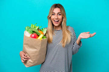 Young Uruguayan woman holding a grocery shopping bag isolated on blue background with shocked...