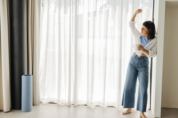 In full growth, slender young caucasian girl, closing eyes, enjoys listening to favorite music. Brunette wears shirt, jeans and drinks morning coffee at home. Comfort concept