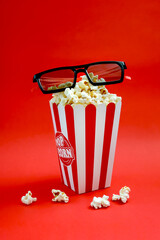 Fresh popcorn in a striped paper cup with 3d glasses on a red background. The concept of cinemas, television, shows. Vertical view