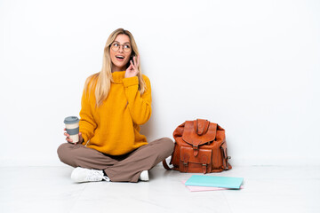 Uruguayan student woman sitting one the floor isolated on white background holding coffee to take away and a mobile