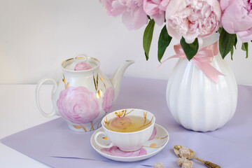 A cup of green tea, a kettle and a vase with pink peonies on the table, lilac background, festive breakfast