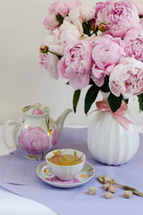 A cup of green tea, a kettle and a vase with pink peonies on the table, lilac background, festive breakfast